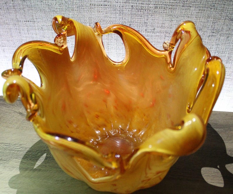May include: A hand-blown glass bowl featuring a vibrant amber and orange swirl pattern. The bowl has a wide, organic, ruffled rim with several distinct, curved glass loops extending upward, creating a dynamic, open, and fluid silhouette.