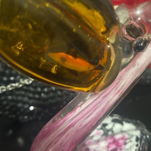 May include: A glass swan figurine featuring a clear neck with pink and white swirling patterns. The body is made of translucent amber-colored glass with a small orange fish-shaped inclusion visible inside the glass structure.