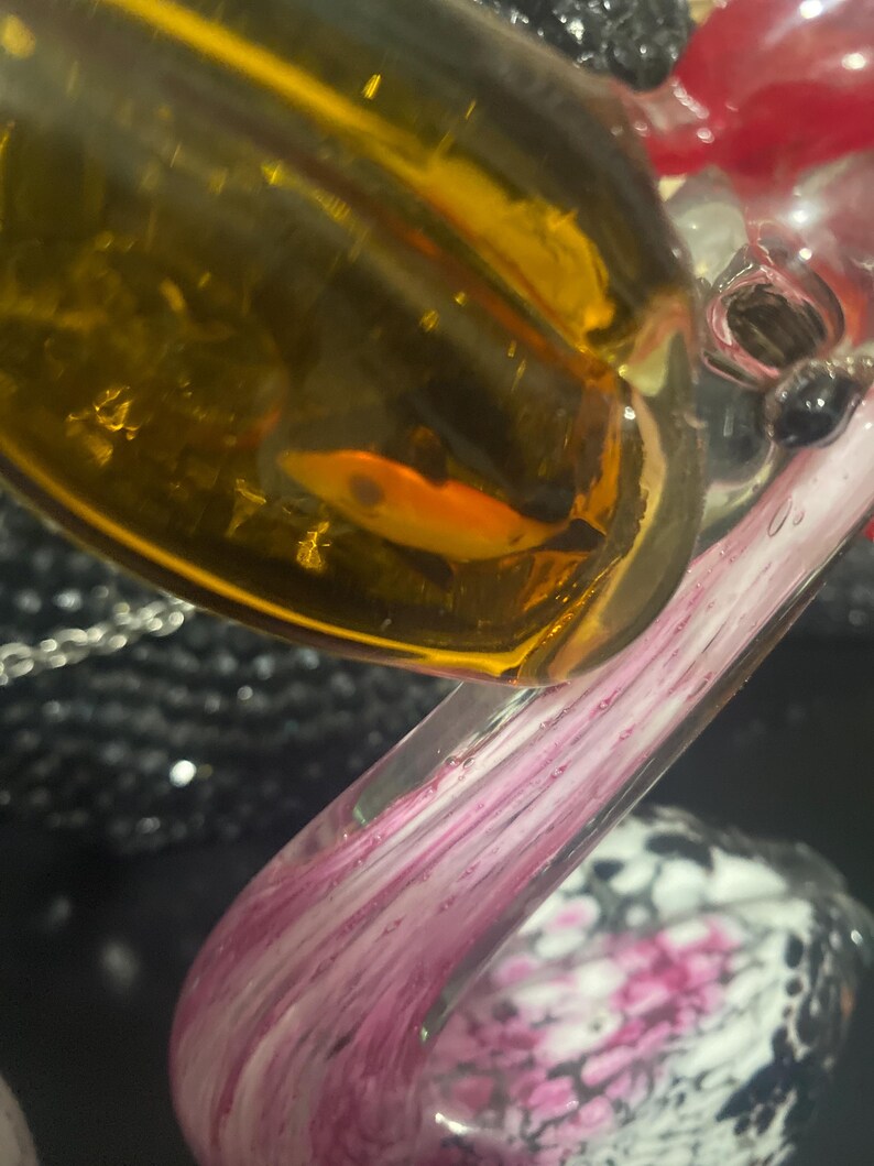 May include: A glass swan figurine featuring a clear neck with pink and white swirling patterns. The body is made of translucent amber-colored glass with a small orange fish-shaped inclusion visible inside the glass structure.