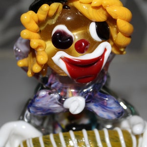 May include: A glass clown figurine with yellow hair, a red nose, and a purple and white patterned shirt. The clown is playing a yellow and white striped accordion.