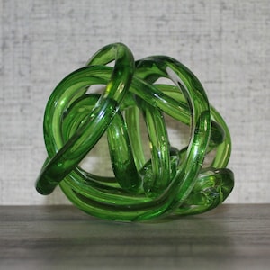 May include: A translucent emerald green glass sculpture formed from a continuous, knotted, and intertwined tube. The object features a smooth, reflective surface with rounded edges, creating a complex, three-dimensional shape with multiple open loops and curves.