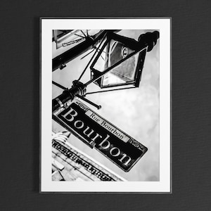 May include: Black and white photo of a street sign in New Orleans that reads "Rue Bourbon" with a vintage street lamp in the background.