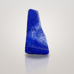 Peut inclure: Une pierre de lapis-lazuli bleu profond, polie et de forme irrégulière. La surface présente des veines blanches et gris clair distinctes qui parcourent le minéral bleu vif, créant un aspect naturel et texturé avec une finition lisse.