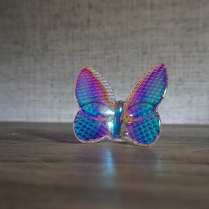 K&ouml;nnte beinhalten: Eine kleine, durchscheinende Schmetterlingsfigur mit einer lebendigen, irisierenden Oberfl&auml;che, die zwischen Blau-, Violett- und Magentat&ouml;nen wechselt. Die Fl&uuml;gel haben eine texturierte Rautenmuster-Oberfl&auml;che, die das Licht einf&auml;ngt und einen reflektierenden, mehrfarbigen Effekt auf dem gesamten Objekt erzeugt.