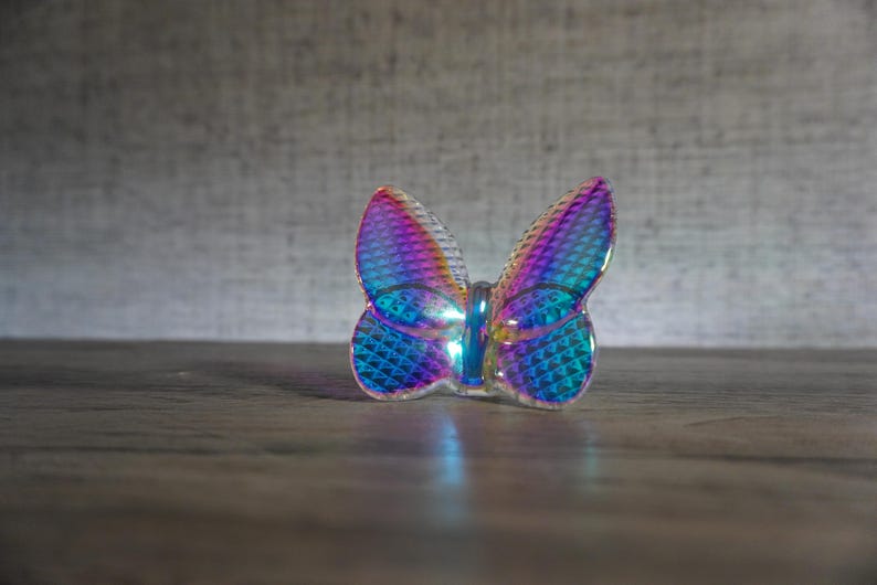 K&ouml;nnte beinhalten: Eine kleine, durchscheinende Schmetterlingsfigur mit einer lebendigen, irisierenden Oberfl&auml;che, die zwischen Blau-, Violett- und Magentat&ouml;nen wechselt. Die Fl&uuml;gel haben eine texturierte Rautenmuster-Oberfl&auml;che, die das Licht einf&auml;ngt und einen reflektierenden, mehrfarbigen Effekt auf dem gesamten Objekt erzeugt.