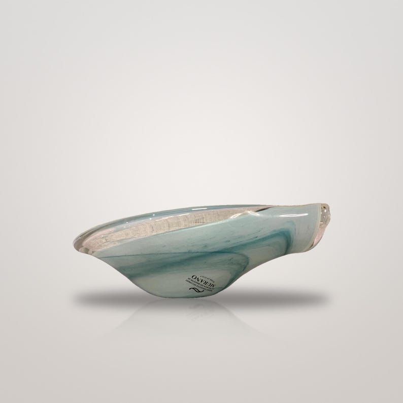 May include: A shallow, asymmetrical glass bowl featuring swirling patterns of translucent light blue and white. The bowl has a smooth, polished finish and includes a small black sticker on the side with the text MURANO.