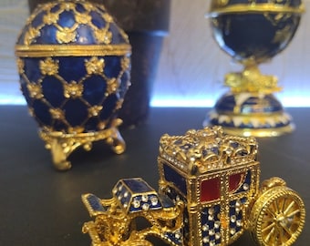 May include: Three gold-plated, jeweled, decorative eggs. One egg is blue with a gold trim and a geometric pattern. Another egg is a blue and gold carriage with red accents. The third egg is a blue and gold globe.