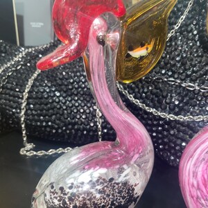 May include: A glass pelican figurine featuring a clear body with pink swirls and black speckled details at the base. The bird has a bright red head and a translucent amber-colored beak containing a small, embedded multi-colored fish graphic.