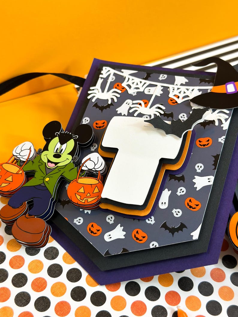 May include: A Halloween-themed banner with a Mickey Mouse character holding a pumpkin bucket. The banner features a black and white spider web design with bats, ghosts, and pumpkins. The banner is layered with different colors of cardstock, including purple, black, and orange.
