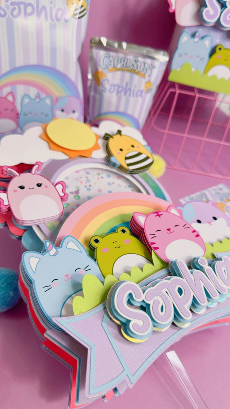 May include: A multi-layered cake topper with a rainbow, clouds, a sun, and a bee. There are also several cartoon animals including a frog, a cat, and a bunny. The topper is decorated with glitter and the name 'Sophia' is written on the bottom.