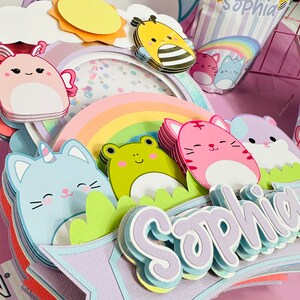 May include: A multi-layered paper craft cake topper with a rainbow, a unicorn cat, a frog, a pink cat, a hamster, a bee, a sun, and a cloud. The topper is decorated with glitter and has the name 'Sophia' written on it. The topper is on a pink background. Capri Sun juice box with the name 'Sophia' on it.