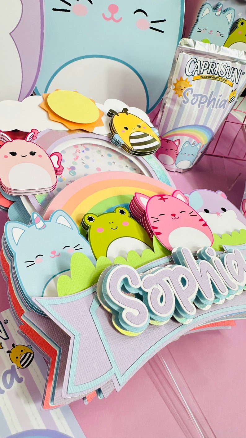 May include: A multi-layered paper craft cake topper with a rainbow, a unicorn cat, a frog, a pink cat, a hamster, a bee, a sun, and a cloud. The topper is decorated with glitter and has the name 'Sophia' written on it. The topper is on a pink background. Capri Sun juice box with the name 'Sophia' on it.