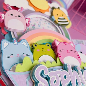 May include: A multi-layered cake topper featuring a rainbow, a sun, a bee, a cat with a unicorn horn, a frog, a cat, and a banner with the name 'Sophia' in purple letters. The topper is made of colorful paper and has a whimsical design.