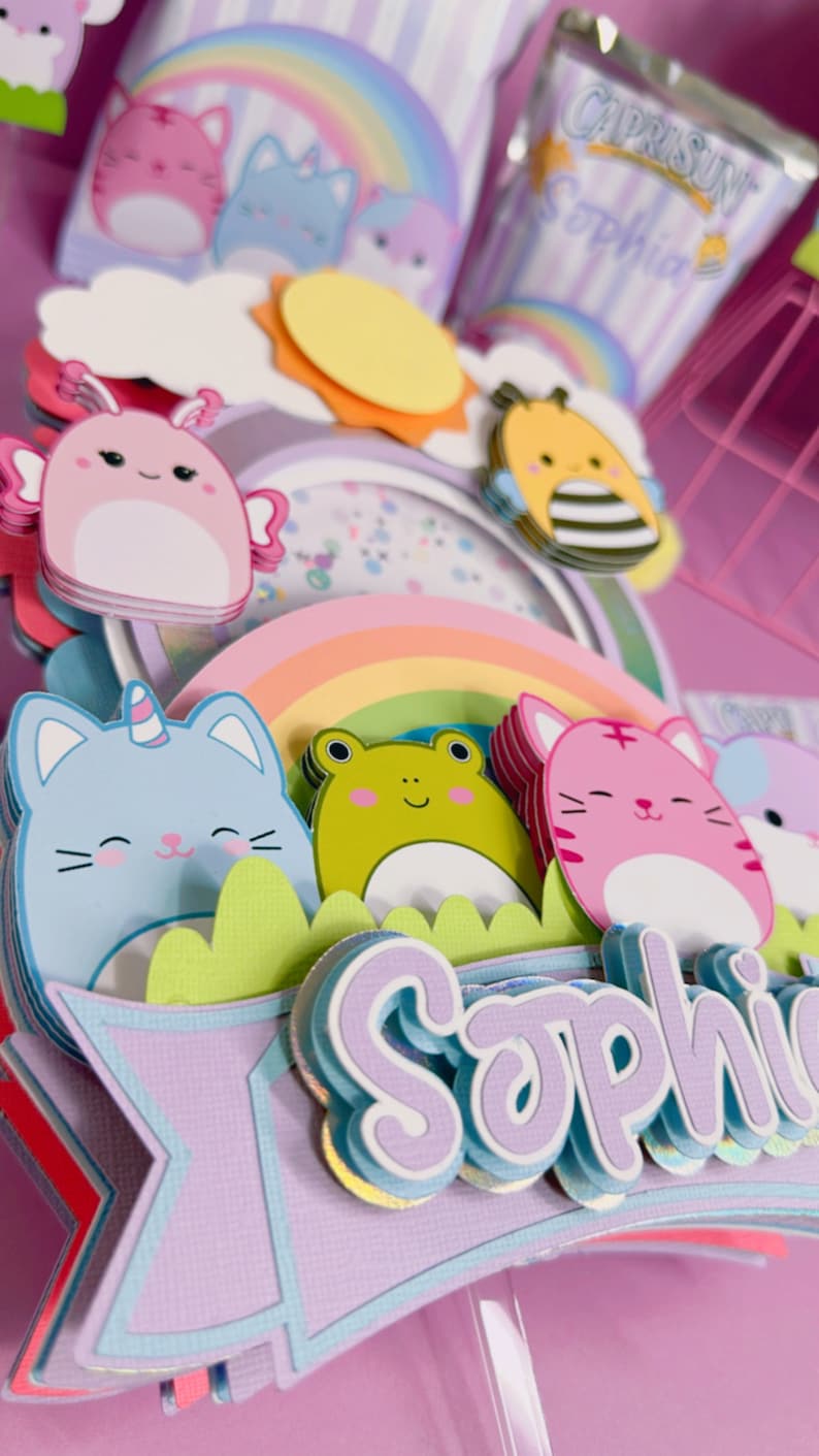 May include: A multi-layered cake topper featuring a rainbow, a sun, a bee, a cat with a unicorn horn, a frog, a cat, and a banner with the name 'Sophia' in purple letters. The topper is made of colorful paper and has a whimsical design.