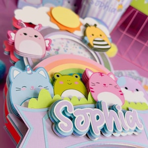 May include: A multi-layered cake topper with a rainbow, a sun, a bee, a frog, and three cats. The topper is decorated with a purple banner that says 'Sophia' in a whimsical font. The topper is on a clear plastic stick.