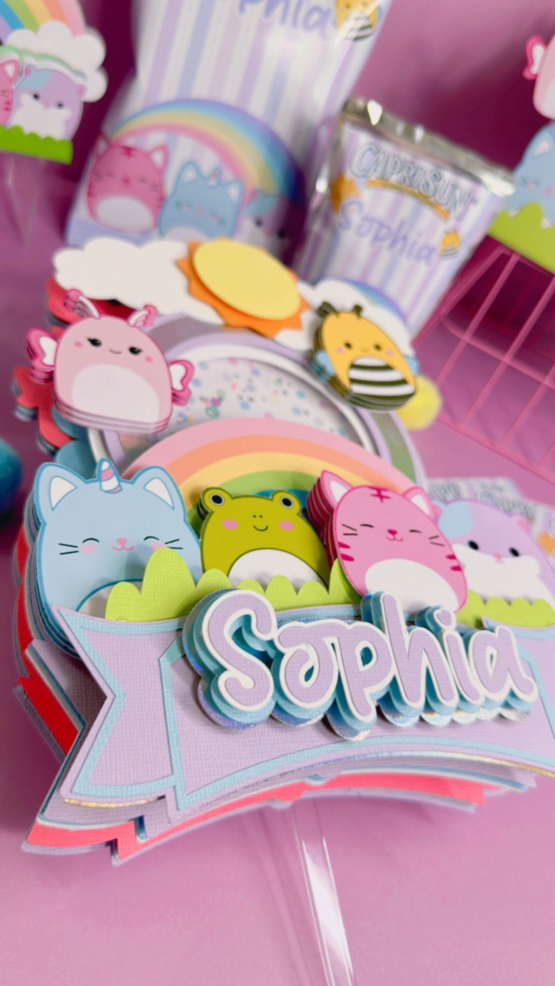 May include: A multi-layered cake topper with a rainbow, a sun, a bee, a frog, and three cats. The topper is decorated with a purple banner that says 'Sophia' in a whimsical font. The topper is on a clear plastic stick.