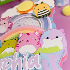 May include: A purple, blue, and pink cake topper with a rainbow, a sun, a bee, a frog, a cat, a hamster, and the name 'Sophia' in a layered font.