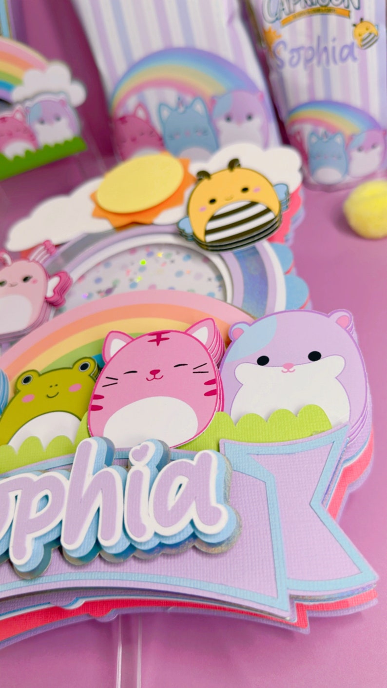 May include: A purple, blue, and pink cake topper with a rainbow, a sun, a bee, a frog, a cat, a hamster, and the name 'Sophia' in a layered font.
