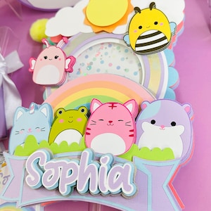 May include: A cake topper with a rainbow, a sun, a bee, a frog, a cat, a hamster, and a butterfly. The topper is decorated with pastel colors and has the name 'Sophia' written on it.