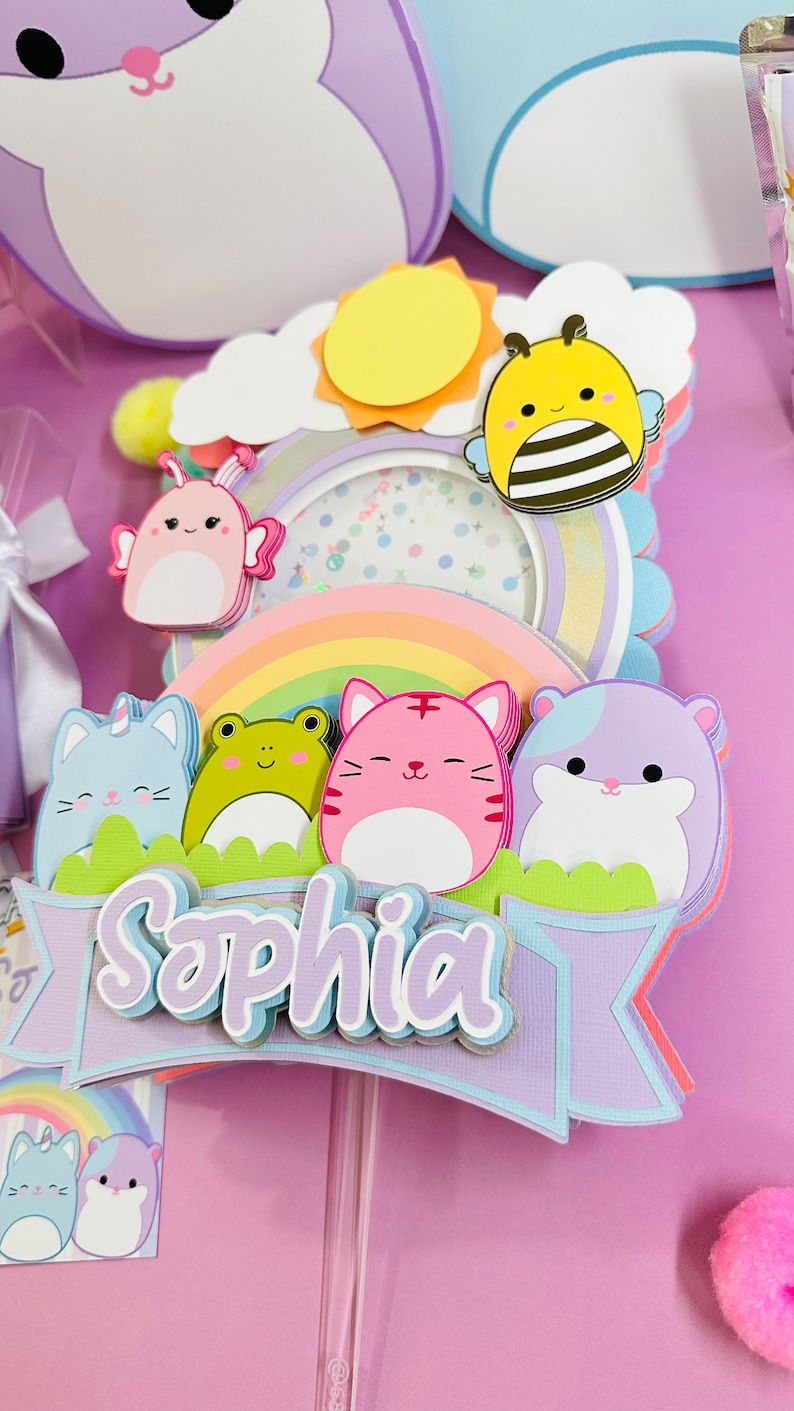 May include: A cake topper with a rainbow, a sun, a bee, a frog, a cat, a hamster, and a butterfly. The topper is decorated with pastel colors and has the name 'Sophia' written on it.