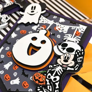 May include: A Halloween-themed banner with a black and white striped background. The banner features a Mickey Mouse character dressed as a skeleton holding a jack-o-lantern. The banner also features a ghost and a spider.