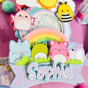 May include: A purple, blue, and green cake topper with a rainbow, a sun, a bee, a frog, a cat, a hamster, and a butterfly. The topper has the name 'Sophia' written on it.