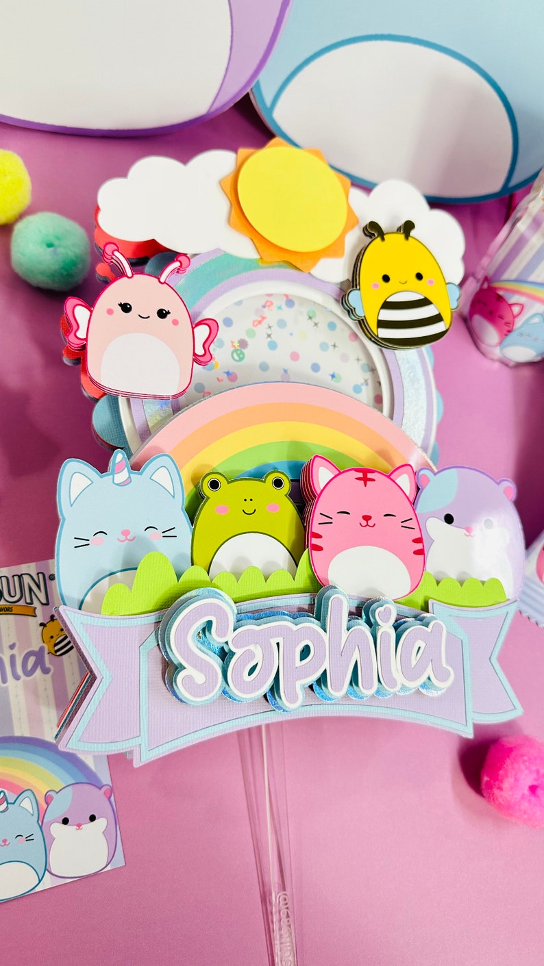 May include: A purple, blue, and green cake topper with a rainbow, a sun, a bee, a frog, a cat, a hamster, and a butterfly. The topper has the name 'Sophia' written on it.
