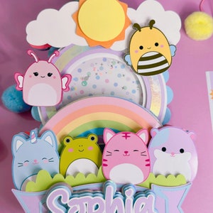 May include: A multi-layered cake topper featuring a rainbow, a sun, a cloud, a bee, a butterfly, a frog, a unicorn cat, a tiger cat, and a hamster. The topper also includes a banner with the name 'Sophia' in purple, blue, and white letters. The topper is made of paper and is decorated with glitter and other embellishments.