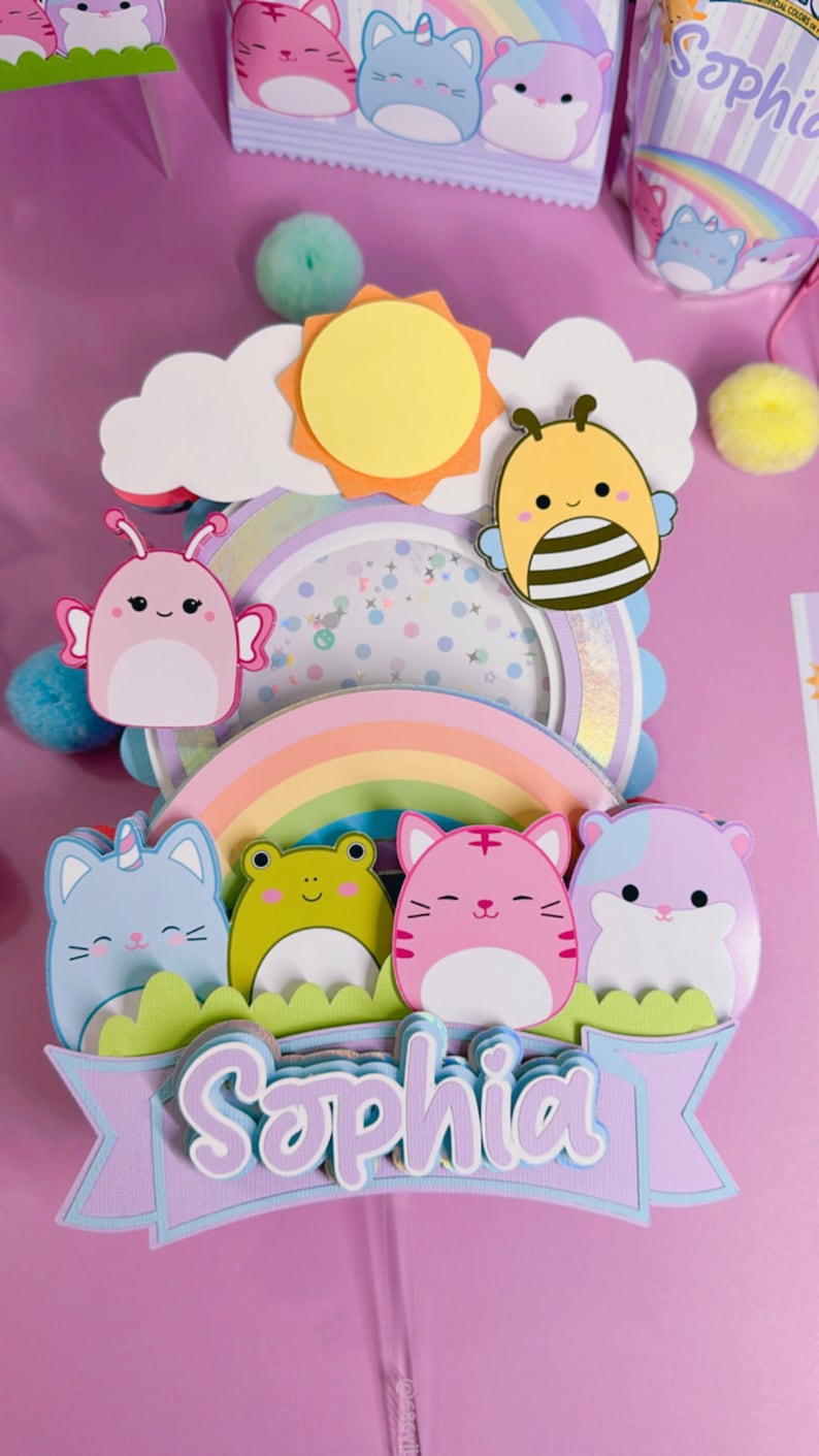 May include: A multi-layered cake topper featuring a rainbow, a sun, a cloud, a bee, a butterfly, a frog, a unicorn cat, a tiger cat, and a hamster. The topper also includes a banner with the name 'Sophia' in purple, blue, and white letters. The topper is made of paper and is decorated with glitter and other embellishments.