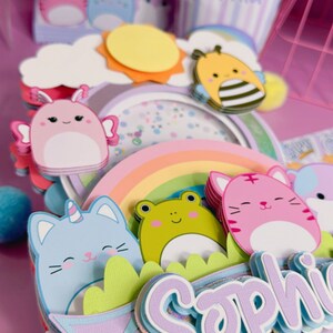 May include: A purple and blue birthday cake topper with a rainbow, a sun, a bee, a frog, a cat, and the name 'Sophia'.