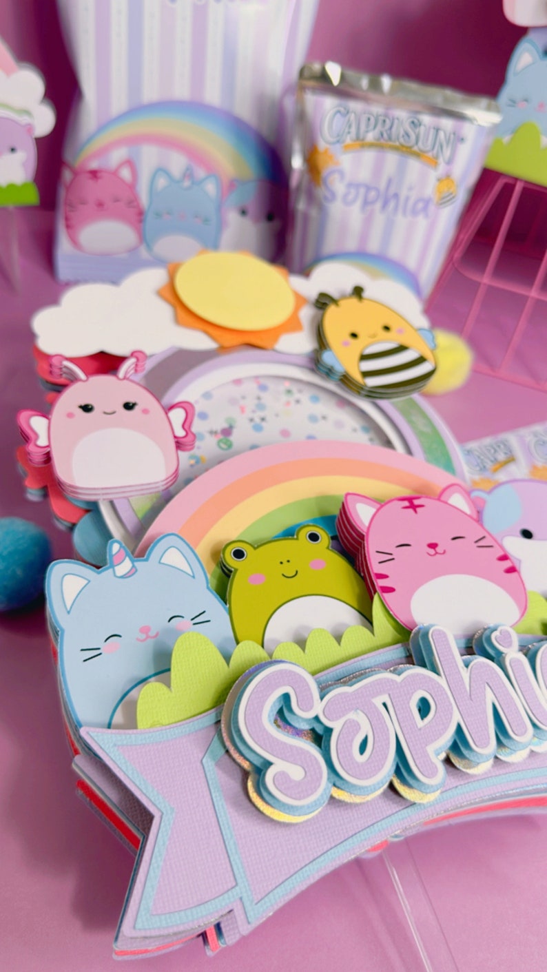 May include: A purple and blue birthday cake topper with a rainbow, a sun, a bee, a frog, a cat, and the name 'Sophia'.