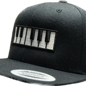 Op de afbeelding: Zwarte snapback cap met een patch met een pianotoetsenbordontwerp in wit en zwart. De cap heeft een platte klep en een klassieke zes-panelen constructie. De hoed is gemaakt van een duurzame stof en is geschikt voor muziekliefhebbers.