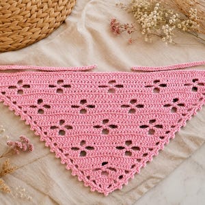 May include: A triangular crochet bandana in a solid light pink colour. The fabric features a repeating openwork pattern of small four-petaled flower shapes. The edges are finished with a delicate picot trim, and two long ties extend from the top corners.