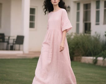 May include: A light pink, floor-length dress with short, flared sleeves. The dress has a round neckline and a gathered waist. The model is walking on a green lawn in front of a white building.