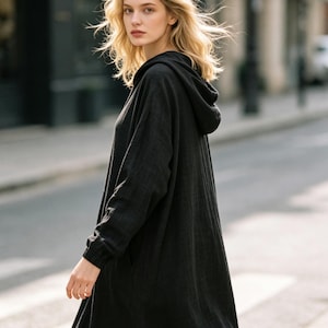 May include: A long-sleeved black hooded dress made of a textured fabric. The garment features a hood, long sleeves with elasticated cuffs, and a loose, flowing silhouette that extends to the lower legs, providing a simple and functional design.