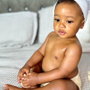 K&ouml;nnte beinhalten: Ein Baby mit einer wei&szlig;en M&uuml;tze sitzt auf einer hellbraunen, strukturierten Decke. Das Baby tr&auml;gt eine Windel und hat dunkelbraunes Haar.
