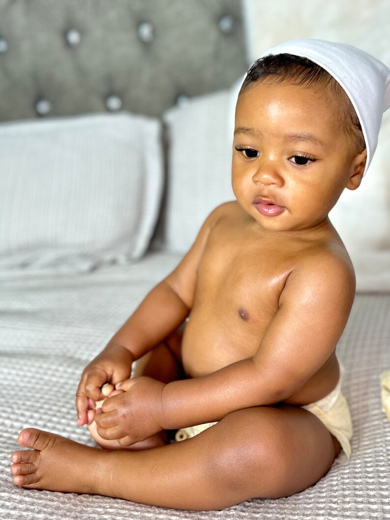 K&ouml;nnte beinhalten: Ein Baby mit einer wei&szlig;en M&uuml;tze sitzt auf einer hellbraunen, strukturierten Decke. Das Baby tr&auml;gt eine Windel und hat dunkelbraunes Haar.