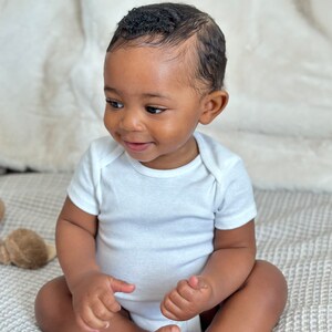 K&ouml;nnte beinhalten: Ein Baby in einem wei&szlig;en Strampler sitzt auf einer strukturierten Decke. Das Baby hat dunkelbraune Haut und kurze, lockige schwarze Haare. Das Baby schaut nach links aus dem Bild.