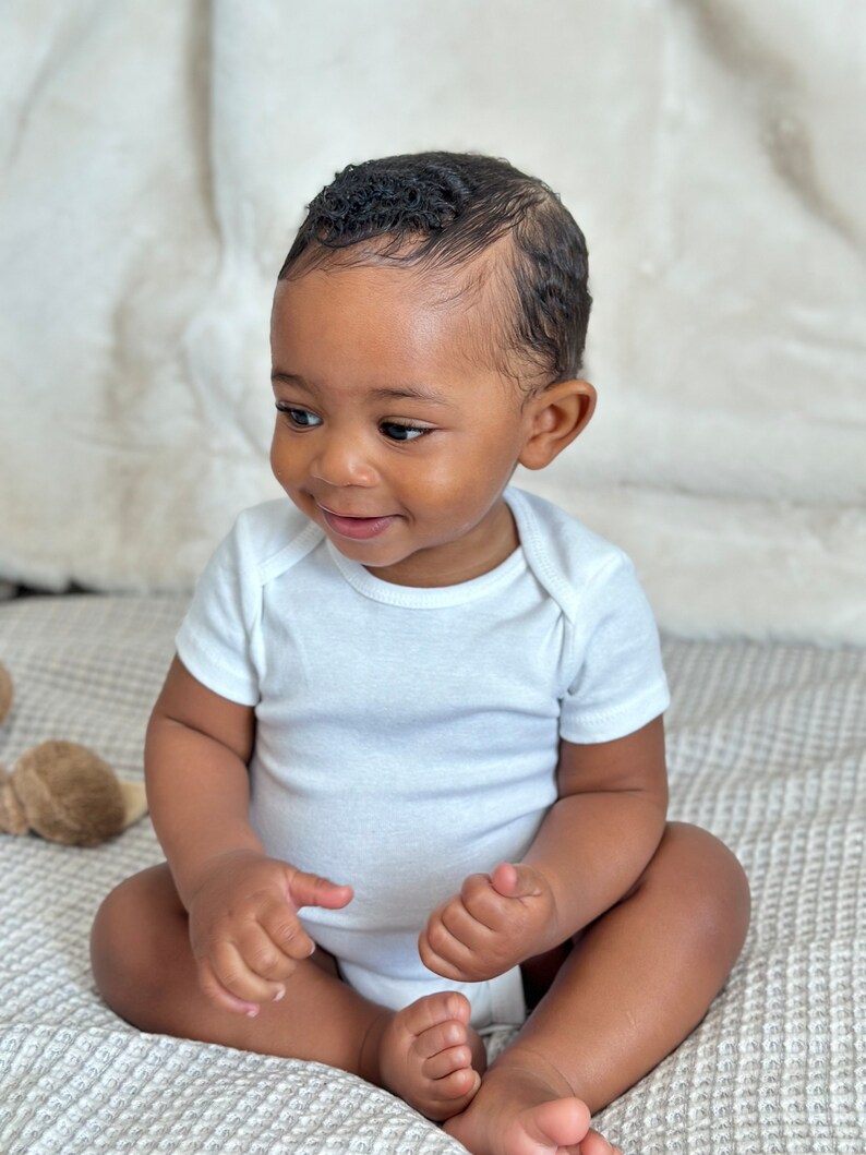 K&ouml;nnte beinhalten: Ein Baby in einem wei&szlig;en Strampler sitzt auf einer strukturierten Decke. Das Baby hat dunkelbraune Haut und kurze, lockige schwarze Haare. Das Baby schaut nach links aus dem Bild.