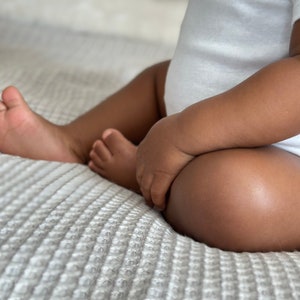 K&ouml;nnte beinhalten: Ein Baby in einem wei&szlig;en Strampler sitzt auf einer wei&szlig;en, strukturierten Decke. Die Beine des Babys sind &uuml;bereinandergeschlagen und die H&auml;nde des Babys ruhen auf seinem Scho&szlig;.