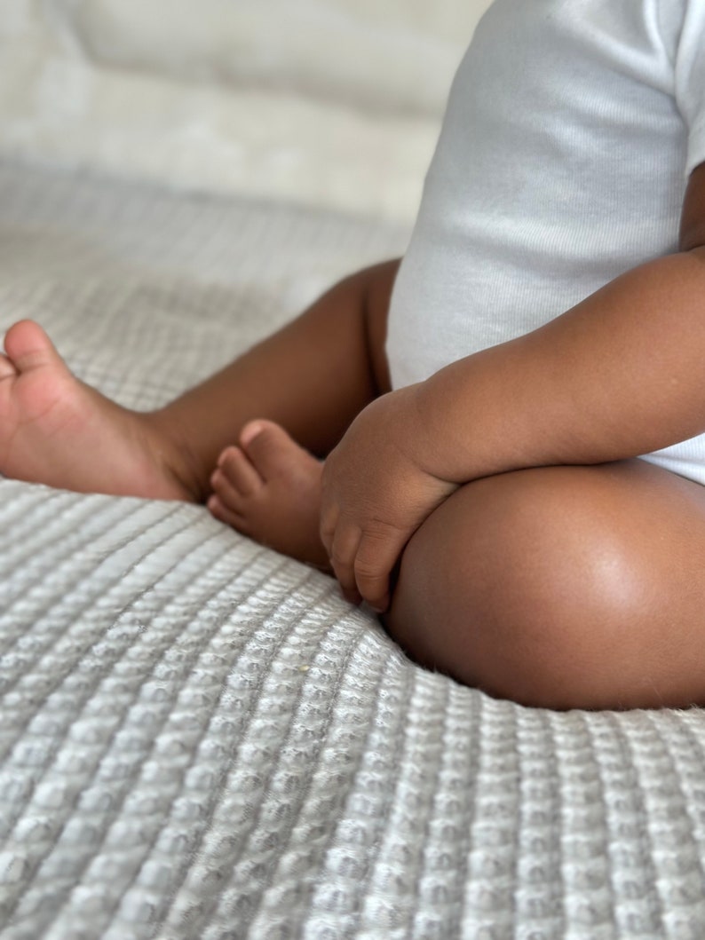 K&ouml;nnte beinhalten: Ein Baby in einem wei&szlig;en Strampler sitzt auf einer wei&szlig;en, strukturierten Decke. Die Beine des Babys sind &uuml;bereinandergeschlagen und die H&auml;nde des Babys ruhen auf seinem Scho&szlig;.