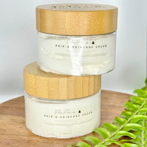 K&ouml;nnte beinhalten: Zwei Glasbeh&auml;lter mit Bambusdeckeln, gef&uuml;llt mit wei&szlig;er Haar- und Hautpflegecreme. Die Beh&auml;lter tragen ein Etikett mit der Aufschrift "PaPa's Hair & Skincare Cream" und einem Kleeblatt-Symbol. Die Beh&auml;lter stehen auf einer Holzoberfl&auml;che mit einer gr&uuml;nen Farnpflanze im Hintergrund.