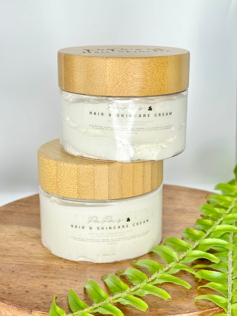 K&ouml;nnte beinhalten: Zwei Glasbeh&auml;lter mit Bambusdeckeln, gef&uuml;llt mit wei&szlig;er Haar- und Hautpflegecreme. Die Beh&auml;lter tragen ein Etikett mit der Aufschrift "PaPa's Hair & Skincare Cream" und einem Kleeblatt-Symbol. Die Beh&auml;lter stehen auf einer Holzoberfl&auml;che mit einer gr&uuml;nen Farnpflanze im Hintergrund.