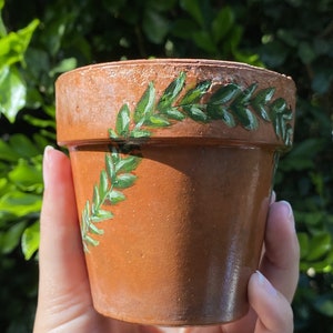 May include: A terracotta flower pot with a hand-painted design of green leaves around the rim.