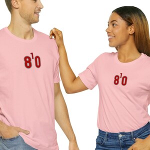 May include: Two people wearing pink t-shirts with the text "8'0" printed on the front. The person on the left is wearing blue jeans. The person on the right is wearing blue jeans.