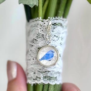 May include: A bouquet of white flowers with green stems, wrapped with white lace. A small, round, silver charm with a blue bird illustration hangs from the bouquet, adding a delicate touch to the floral arrangement.