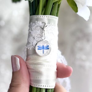 May include: A bouquet of white flowers and green stems, wrapped with white ribbon and lace. A silver charm with a blue dragonfly design is attached to the ribbon, adding a delicate touch. The charm is round and has a clear background.