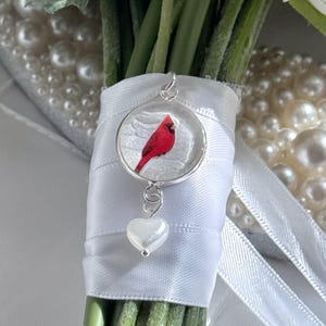 May include: A silver pendant with a red cardinal bird design, suspended from a white ribbon-wrapped bouquet. The pendant has a heart-shaped charm. The bouquet includes white flowers and green stems, with a pearl backdrop.