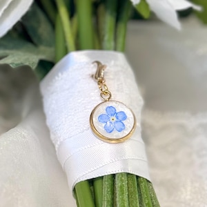 May include: A gold-colored charm with a blue flower design, attached to a white ribbon-wrapped bouquet. The charm features a small, round pendant with a delicate blue floral pattern set against a white background. The bouquet has green stems and white flowers.