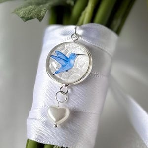 May include: A silver pendant with a blue hummingbird design and a white heart charm is attached to a bouquet of white flowers wrapped in white ribbon. The hummingbird is depicted in flight, with its wings spread.